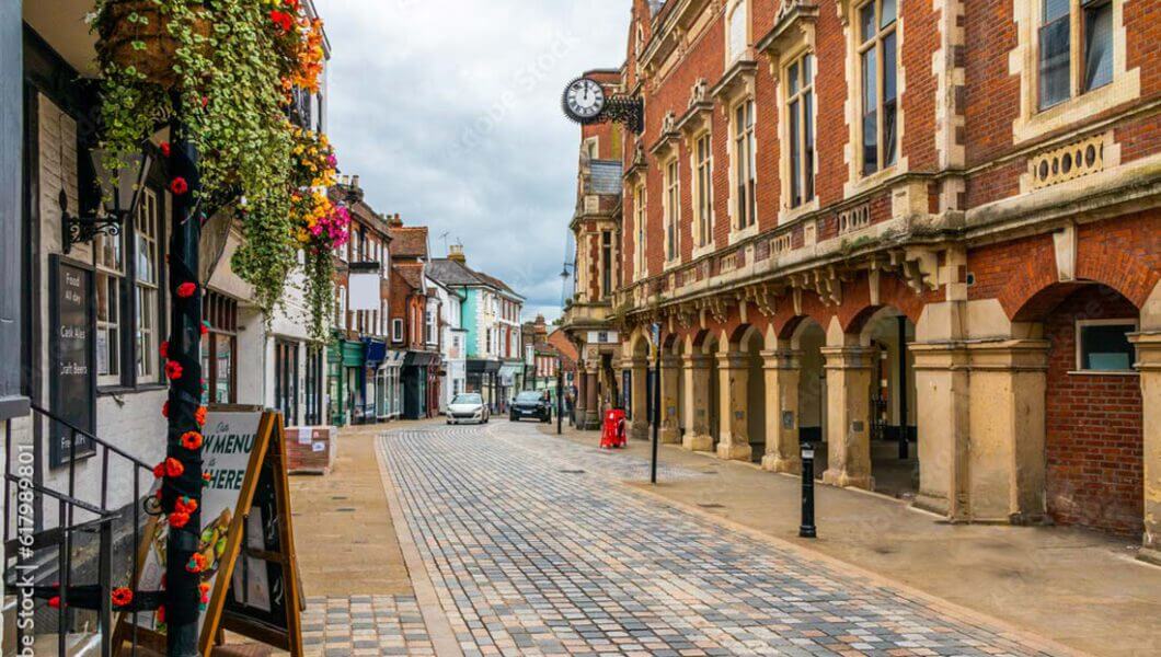 A quaint cobblestone street lined with historic buildings and colorful hanging flowers. A clock is mounted on one of the buildings. A restaurant sign on the left reads "MENU HERE." The scene is peaceful with no visible pedestrians or vehicles.