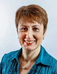 A woman with short brown hair smiles at the camera. She is wearing a teal blouse with a textured pattern and a small necklace. The background is a plain, light color.