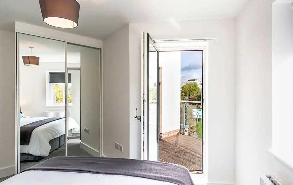 A modern bedroom with white walls features a bed with a white duvet and gray throw. A mirrored wardrobe reflects the room. Next to the bed, a glass door opens to a small balcony with outdoor views. The room is bright with natural light.