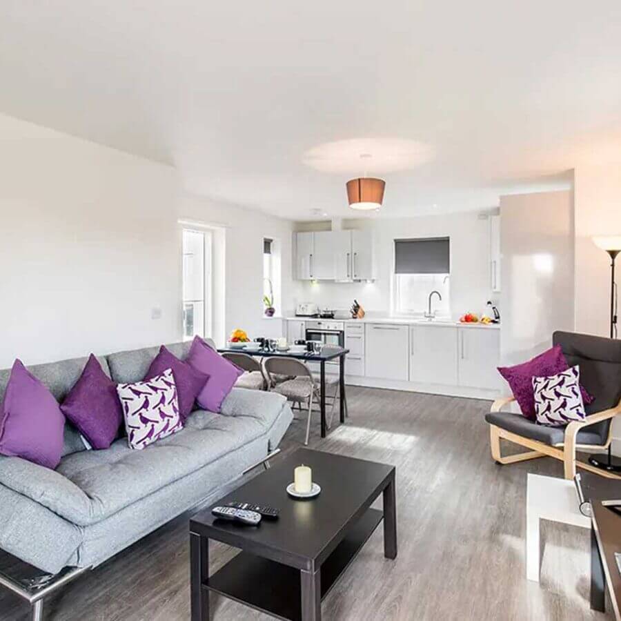 A modern open-plan living area with a gray sofa adorned with purple and white patterned pillows, a black coffee table, and a flat-screen TV. A kitchen with white cabinets and stainless steel appliances is visible in the background. The room has wooden flooring.