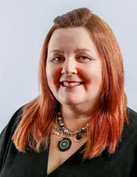 A woman with shoulder-length reddish hair, smiling, wearing a black top and a decorative necklace with a large pendant. She is against a plain gray background.