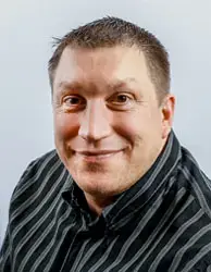 A man with short, brown hair and a friendly smile is facing the camera. He is wearing a black, collared shirt with white vertical stripes. The background is plain and light-colored.
