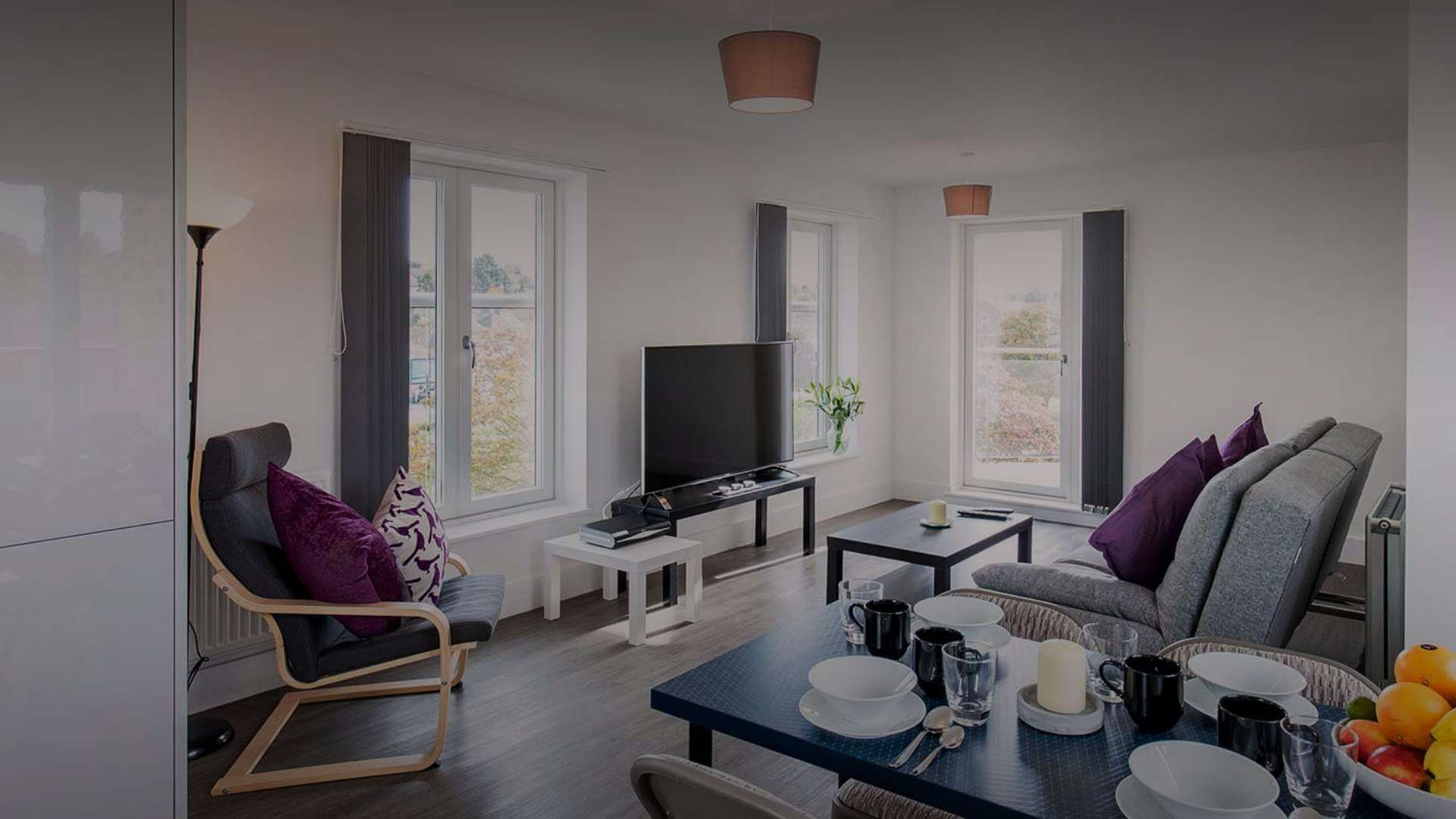 A modern living and dining area with large windows, a TV, a coffee table, and a couch with purple pillows. A dining table is set with plates, mugs, and a candle centerpiece. There's a wooden chair with a cushion and a standing lamp in the corner.