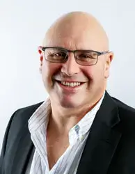 A bald man wearing glasses, a white shirt, and a black blazer is smiling at the camera. The background is plain and light-colored.