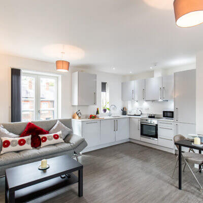 A modern, open-plan living space featuring a kitchen and a cozy seating area. The kitchen has white cabinets and stainless steel appliances. The living area includes a gray sofa with red and white pillows, a black coffee table, and a small dining table with two chairs.
