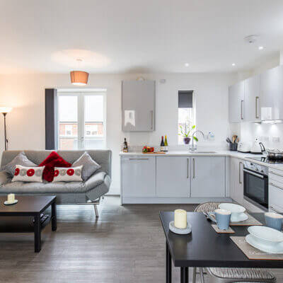 A modern, open-concept living room and kitchen area. The space features a gray couch with red and gray pillows, a black coffee table, and a small dining table set for two. The kitchen has sleek gray cabinets, appliances, and a countertop with fruit and decor.