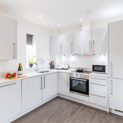 A modern white kitchen with grey countertops, featuring a built-in oven, stove, and stainless steel sink. There are white cabinets, a row of knives in a wooden block, a chopping board with vegetables, and a bottle of wine on the counter.