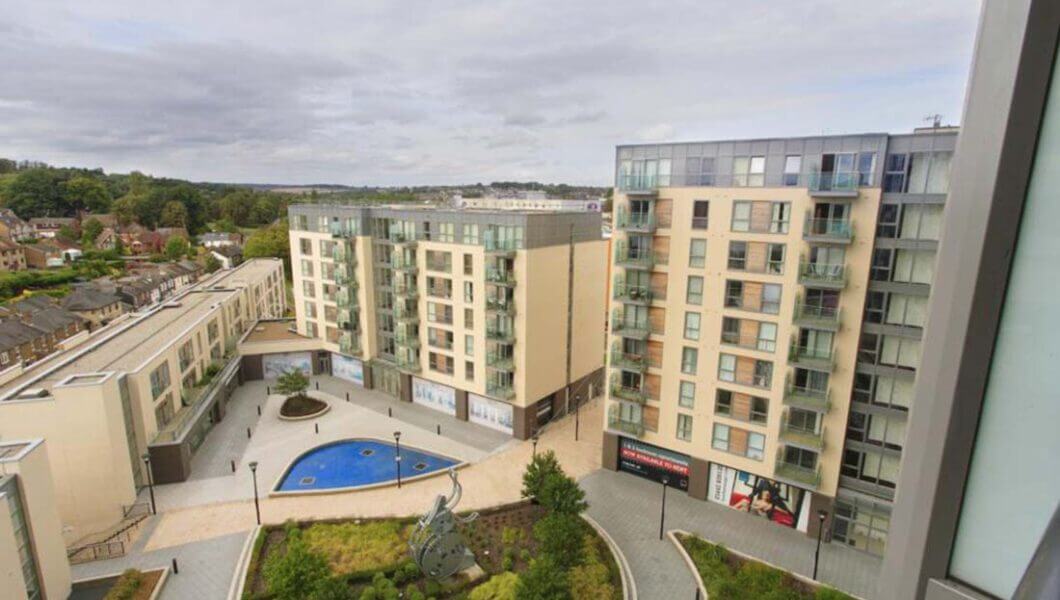 Aerial view of a modern apartment complex featuring tall buildings with balconies, a courtyard with green landscaping, a small fountain, and surrounding walkways. Residential houses and a wooded area can be seen in the background under a cloudy sky.