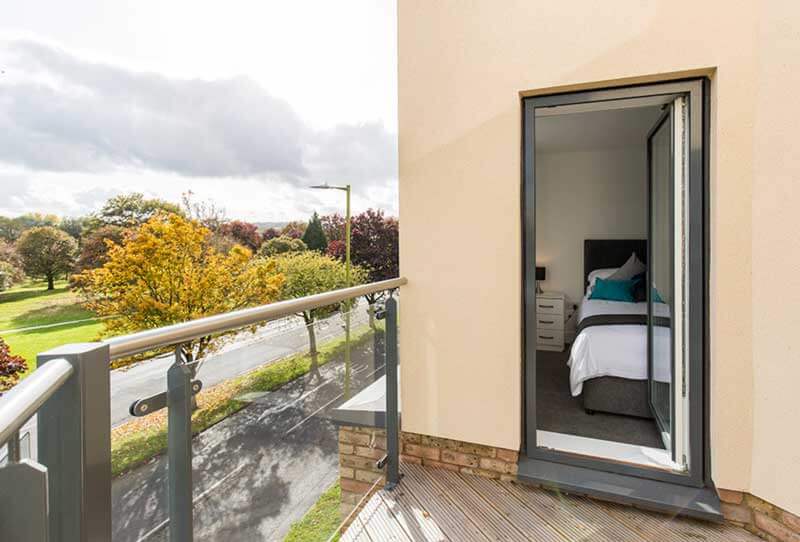 A modern bedroom with white walls features a bed with a white duvet and gray throw. A mirrored wardrobe reflects the room. Next to the bed, a glass door opens to a small balcony with outdoor views. The room is bright with natural light.