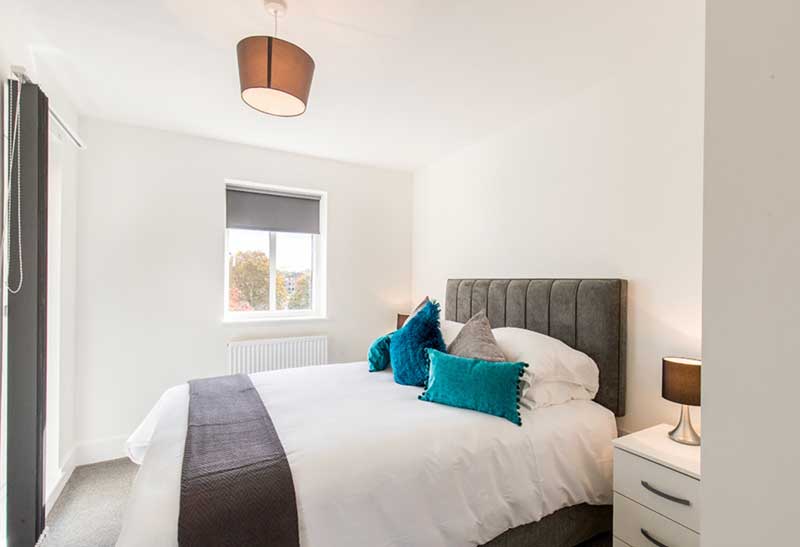 A bedroom with a neatly made double bed featuring a white duvet, gray headboard, and teal accent pillows. A small bedside table with a lamp is on the right. There is a window with a gray blind, a ceiling light, and the room is decorated in white and gray tones.