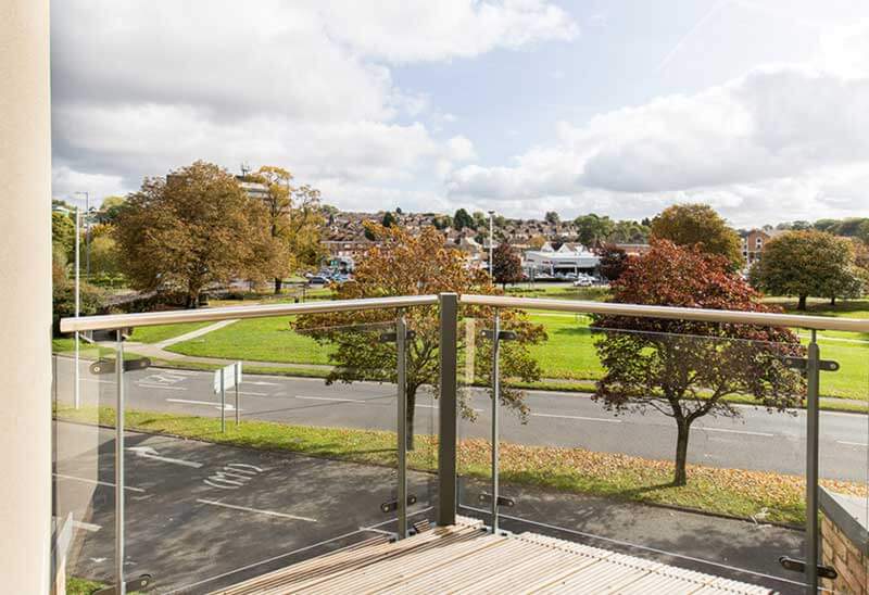 A view from a balcony with a glass railing overlooking a peaceful neighborhood park. The park has green grass and several trees with autumn foliage. In the background, there are houses and a hill covered with trees. The sky is partly cloudy.