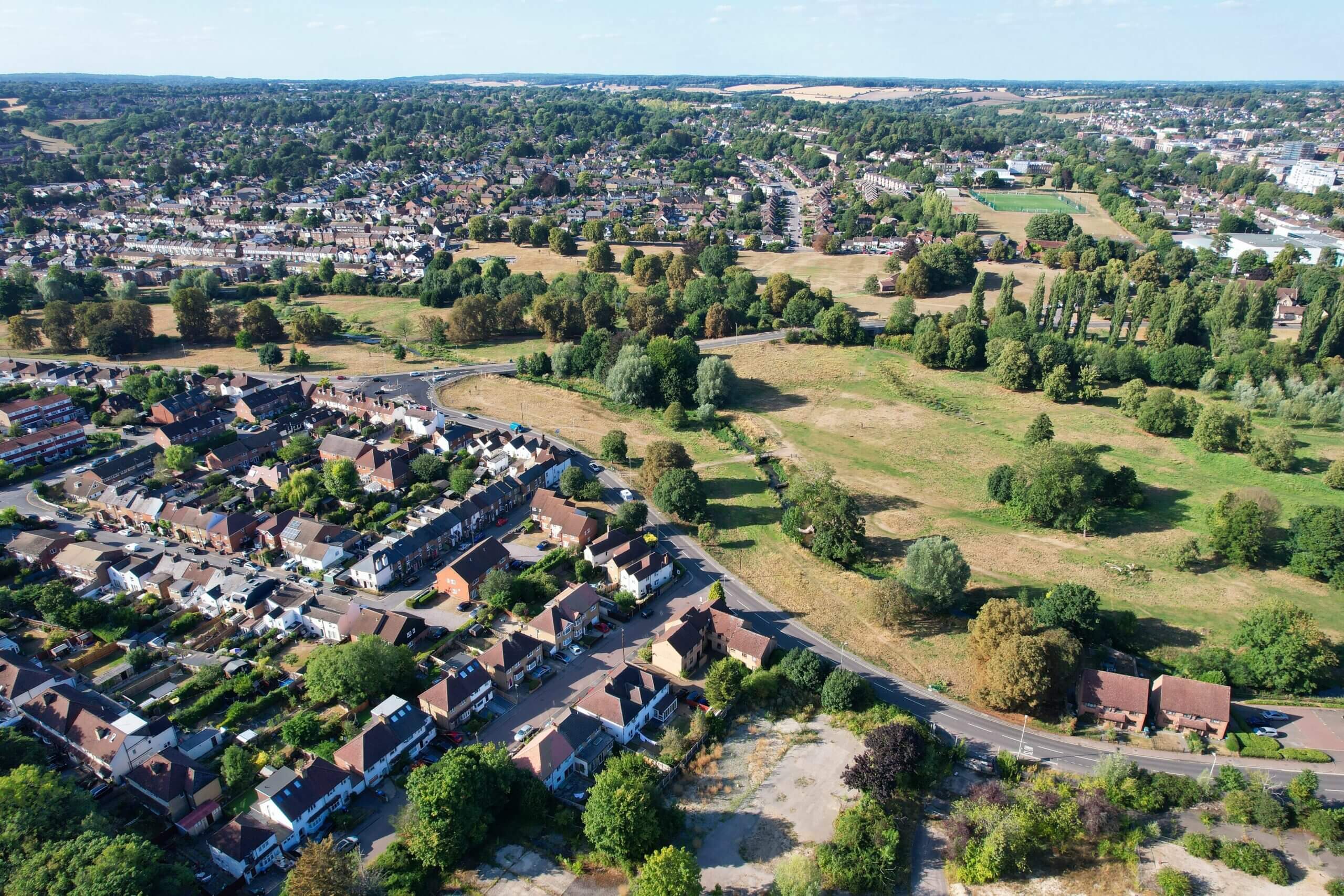 An aerial view of a suburban neighborhood and green open fields. The left side features rows of houses with adjacent roads, while the right side showcases expansive grasslands interspersed with trees. The background includes more greenery and additional housing.