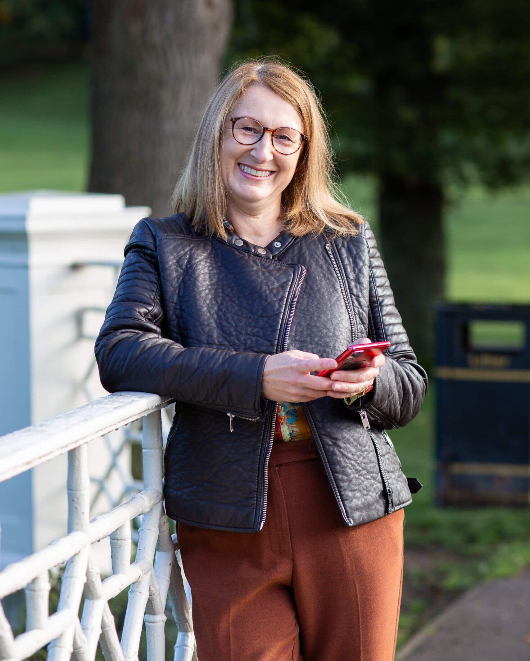 A woman with shoulder-length brown hair smiles at the camera. She is wearing a brown jacket and holding a moble phone in her hand and is wearing glasses. The background is trees in a park.